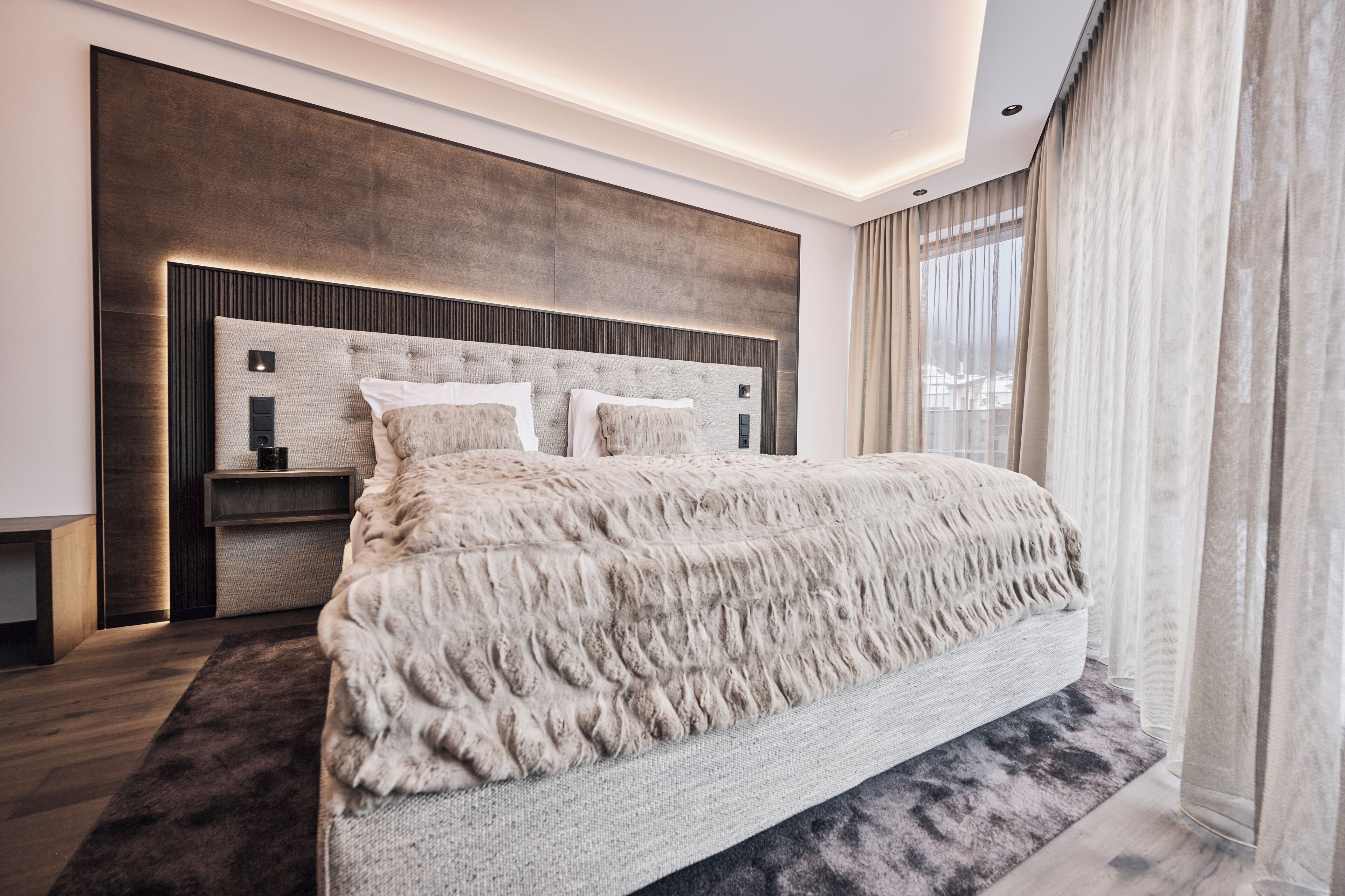 Ein modernes Schlafzimmer mit einem großen Bett, einem Kopfteil mit integrierter Beleuchtung, einem Nachttisch und einem großen Fenster mit Vorhängen.