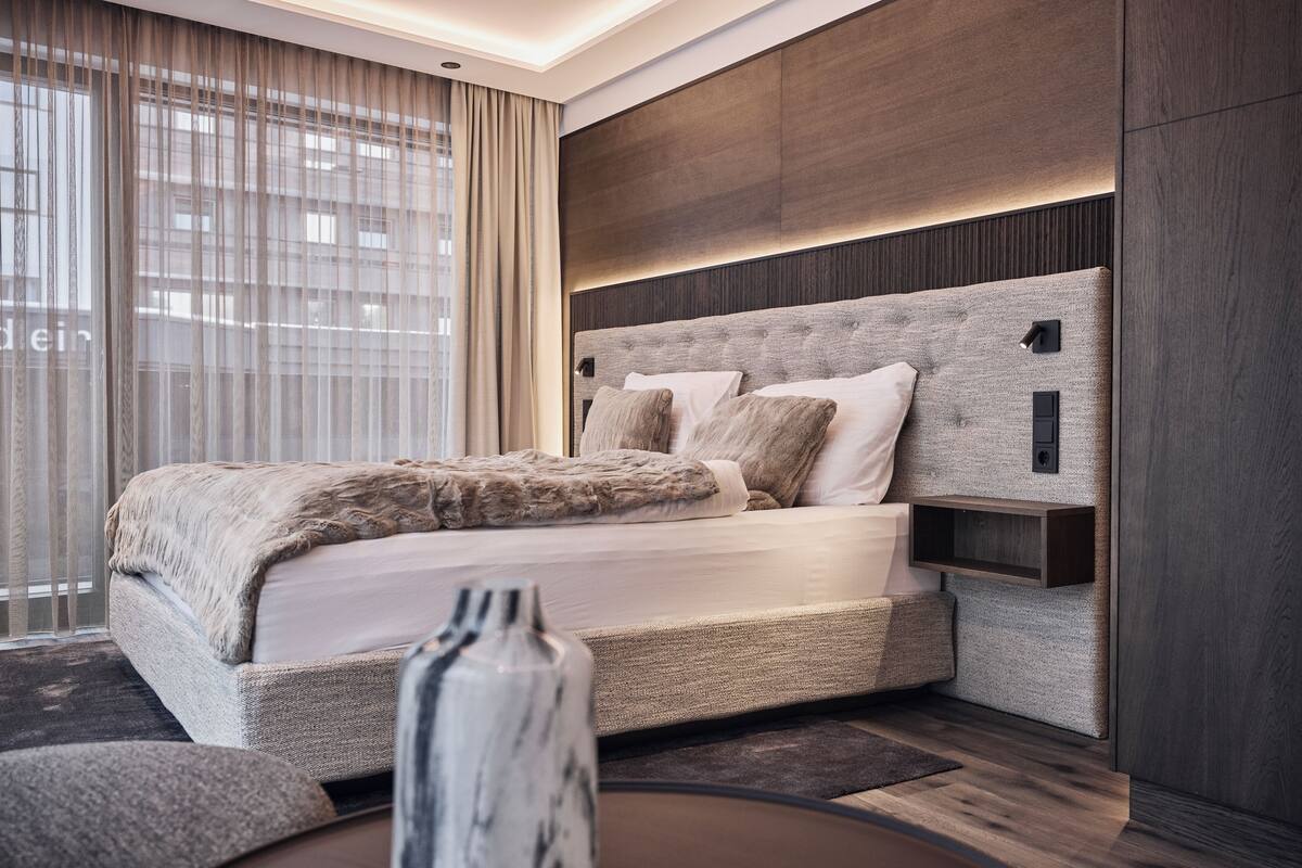 Ein modernes Schlafzimmer mit einem großen Bett, einem an der Wand befestigten Kopfteil mit integrierter Beleuchtung, einem Nachttisch und einem großen Fenster mit Vorhängen.