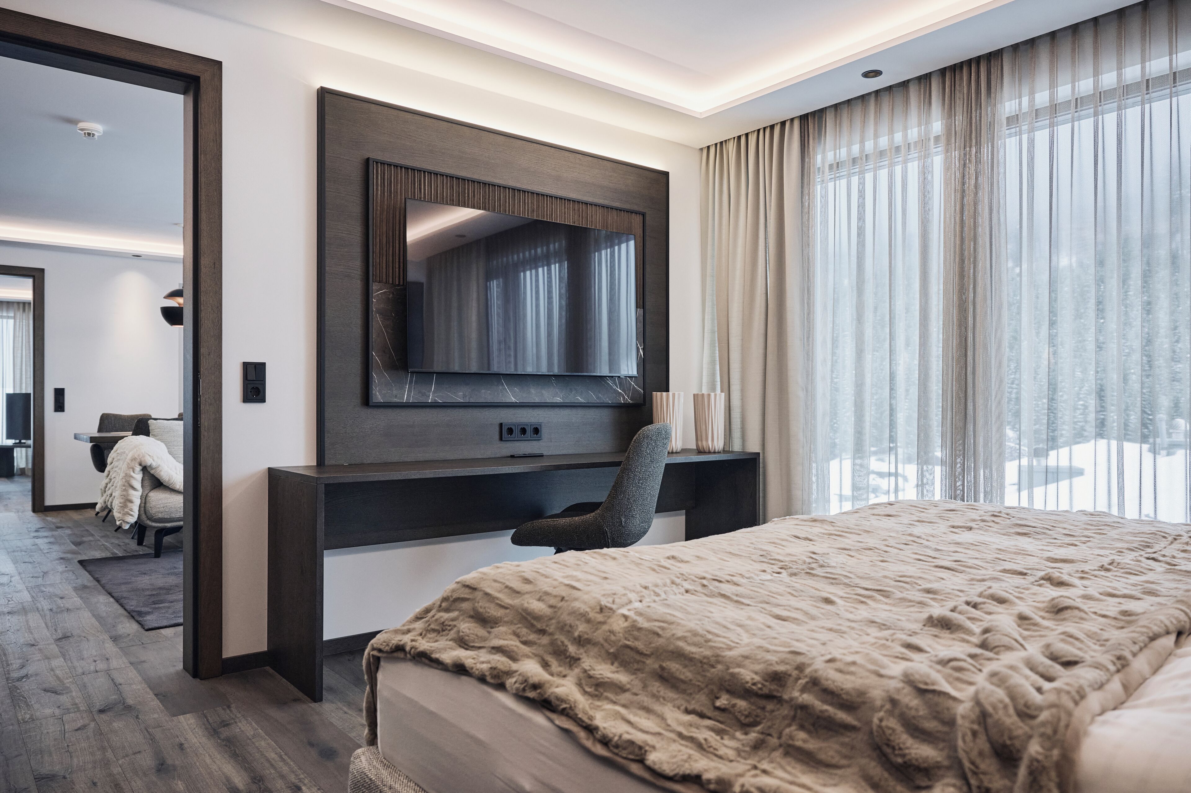 Moderni hotellihuone, jossa on suuri sänky, seinään kiinnitetty työpöytä tuolilla, suuri peili ja näkymä lumiseen maisemaan ikkunasta.