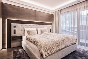 A modern bedroom with a large bed, a headboard with integrated lighting, a bedside table, and a view of a balcony through sheer curtains.