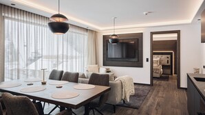 A modern hotel room with a dining area, a large window with curtains, a flat-screen TV mounted on the wall, and a cozy seating area with a sofa and a chair. - Chalet Ischgl - Modern Luxury Residences (Ischgl)