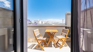 Een balkon met twee houten stoelen en een tafel, een heldere blauwe lucht en gebouwen op de achtergrond.