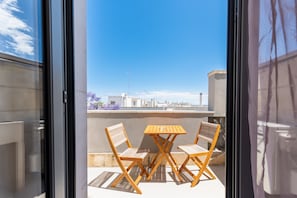 A balcony with two wooden chairs and a table, a clear blue sky, and buildings in the background.