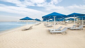 Ein Strand mit Liegestühlen und blauen Sonnenschirmen, bewölkter Himmel und ruhigem Meer.