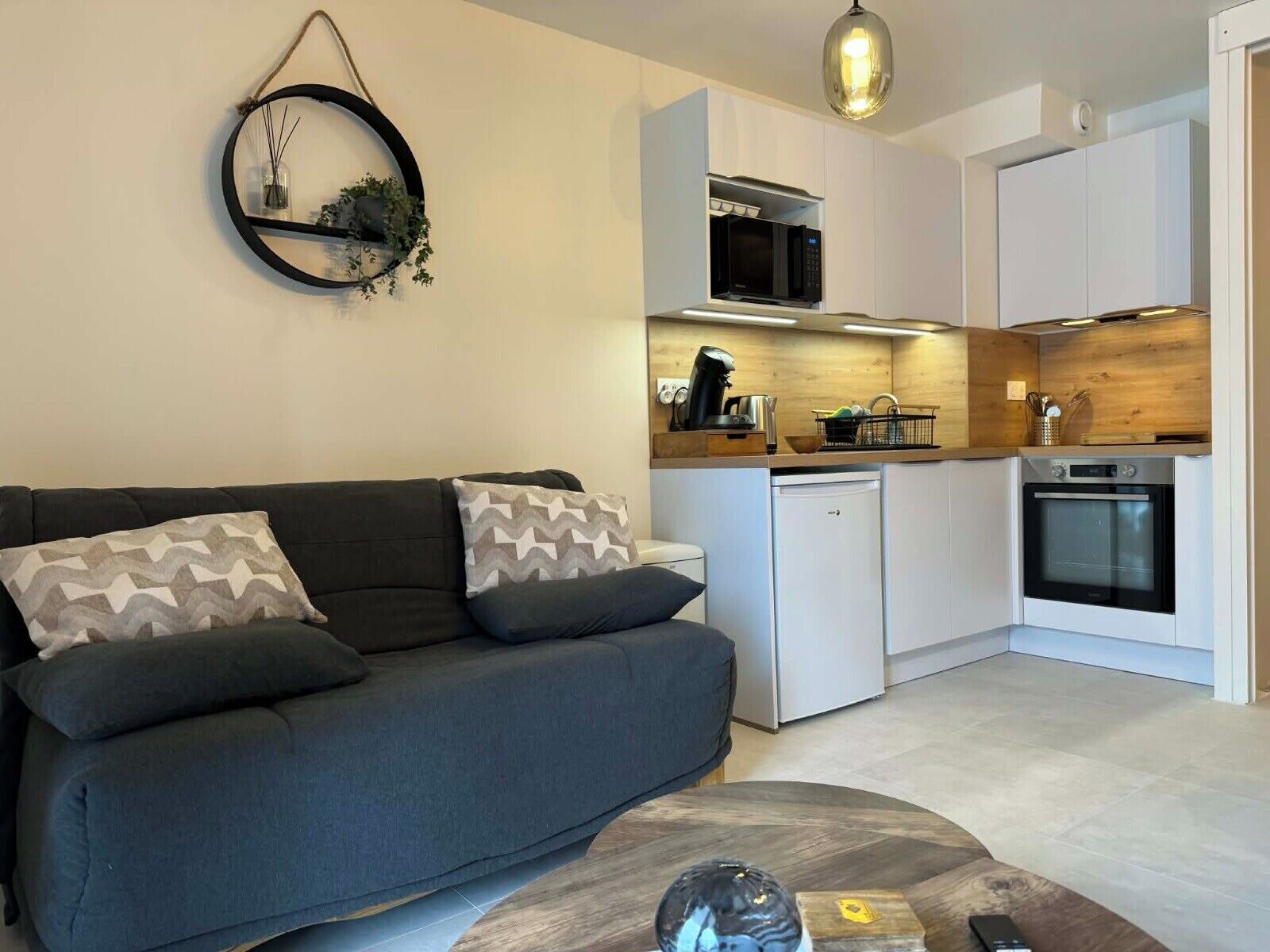 Een moderne keuken met witte kasten, een ingebouwde magnetron en een oven. Er staat een grijze bank met decoratieve kussens en een klein bijzettafeltje met een decoratie.