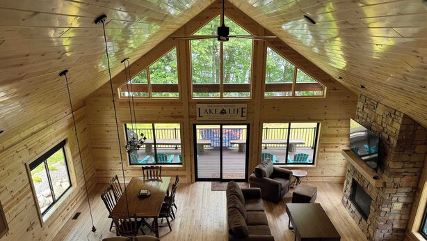 Une cabane spacieuse avec un plafond haut, des murs en bois, un coin repas, un salon avec des canapés, une cheminée en pierre et de grandes fenêtres offrant une vue sur l’extérieur.