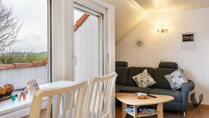 A cozy room with a sofa, a small table, and a window overlooking a garden. - Meeresfeeling for 2: Cozy North Sea Apartment, 34 sqm, 1 Bedroom, Max 2 Guests (Horumersiel)