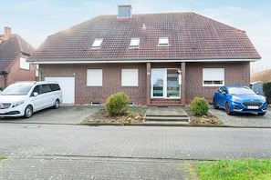 Une maison à deux étages, construite en briques, avec un toit recouvert de tuiles, deux petits arbustes et deux voitures garées.