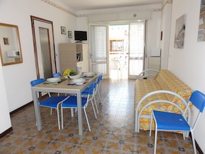 Un coin repas avec une table dressée pour quatre personnes, des chaises bleues, un canapé avec des coussins à motifs et un téléviseur posé sur un meuble.