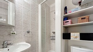 A bathroom with a sink, a glass shower enclosure, and a toilet. There are shelves with toiletries and a mirror above the sink. - Bright apartment near beach (Narbonne)