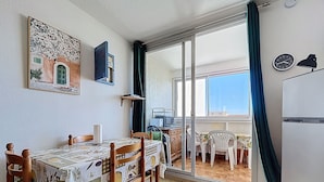 A dining area with a table set for four, wooden chairs, a patterned tablecloth, a wall-mounted clock, and a view through a glass door to an outdoor balcony. - Bright apartment near beach (Narbonne)