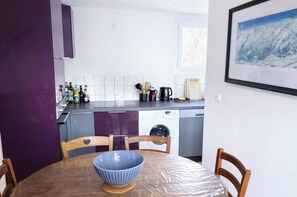 A modern kitchen with a dishwasher, a coffee maker, and a window. There is a table with a blue bowl and chairs.