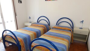 A room with two beds, each with a blue metal frame and colorful striped bedding. There are bedside tables with lamps and a small shelf. - Apartment Close to the Thermal Baths (San Michele al Tagliamento)