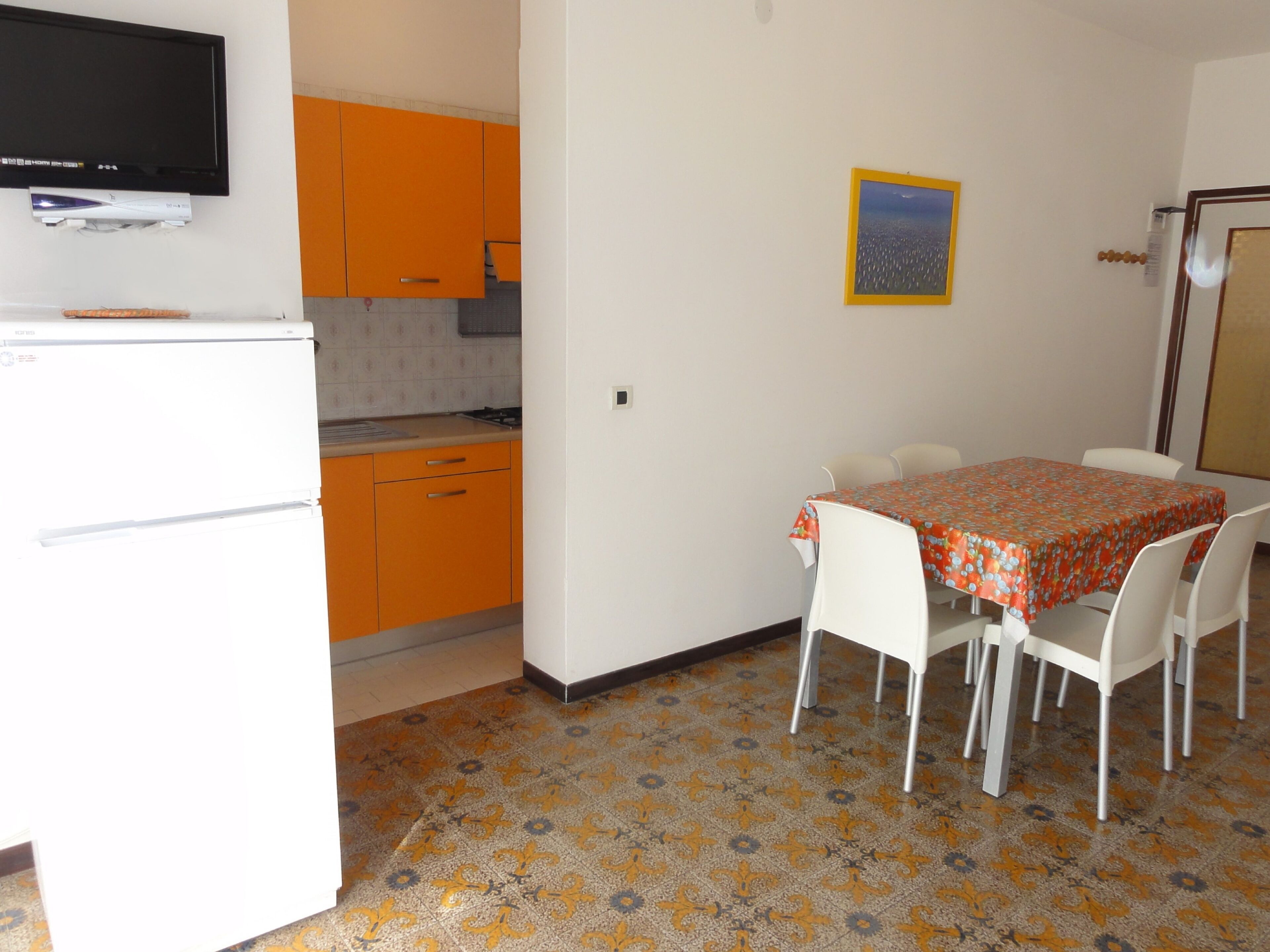 Una cucina compatta con pensili arancioni, un frigorifero bianco e un'area pranzo con un tavolo ricoperto da una tovaglia fantasia.