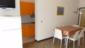 A compact kitchen with orange cabinets, a white refrigerator, and a dining area with a table covered in a patterned tablecloth. - Apartment Close to the Thermal Baths (San Michele al Tagliamento)