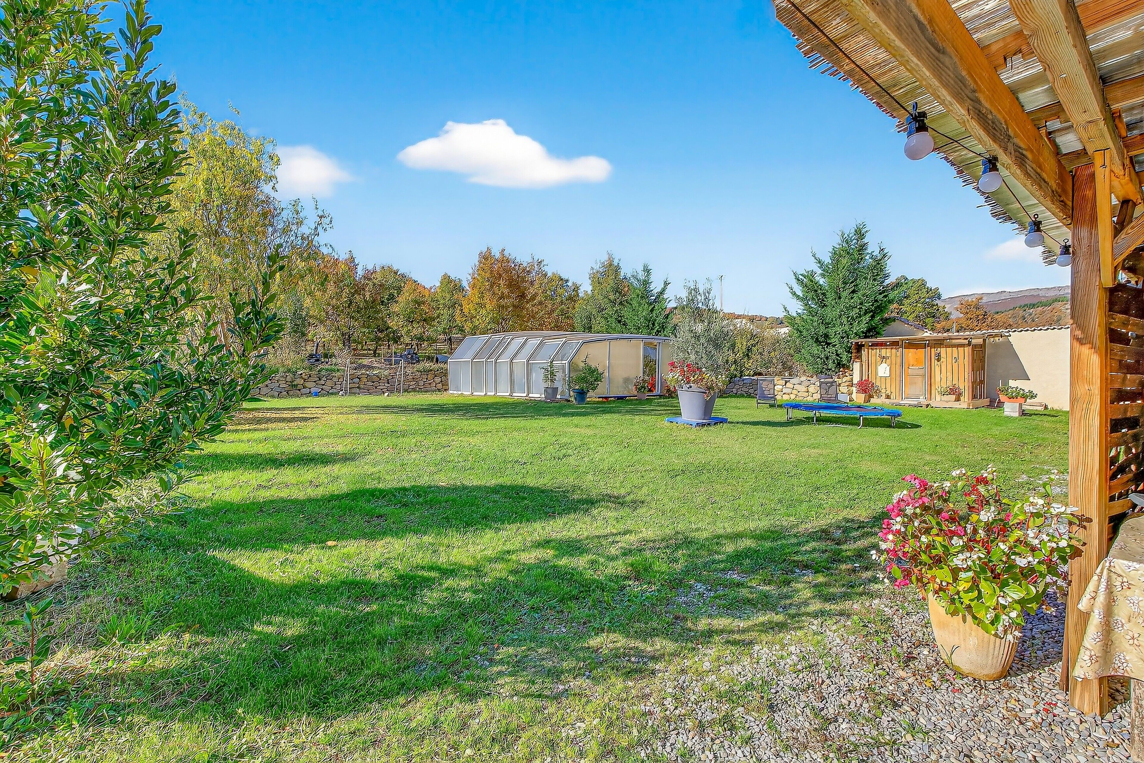 A backyard with a wooden pergola, a garden with various plants, a shed, and a small greenhouse.