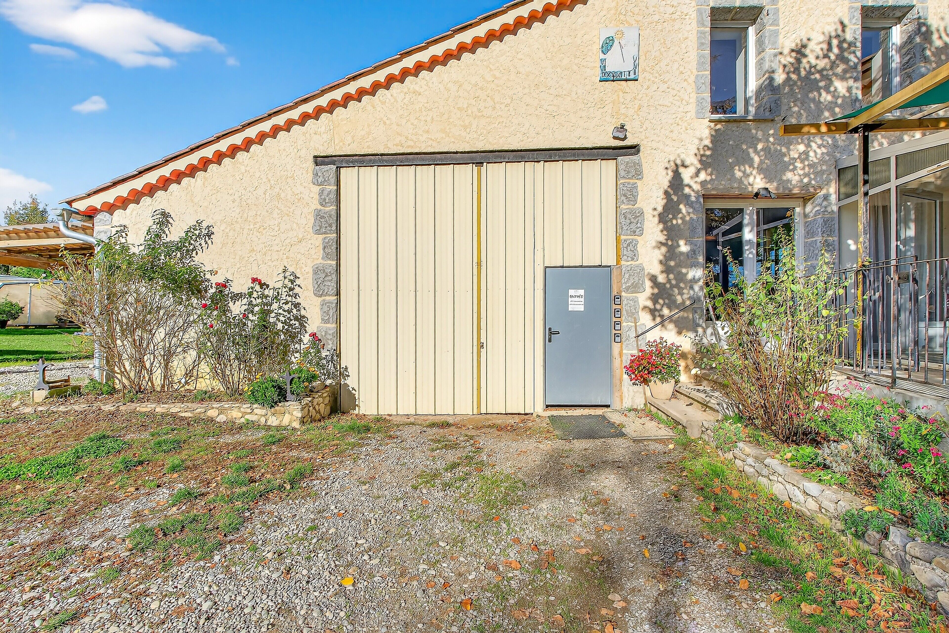 Un bâtiment à la façade beige, avec un grand portail de garage et une petite porte. On y trouve des plantes et une allée gravillonnée.