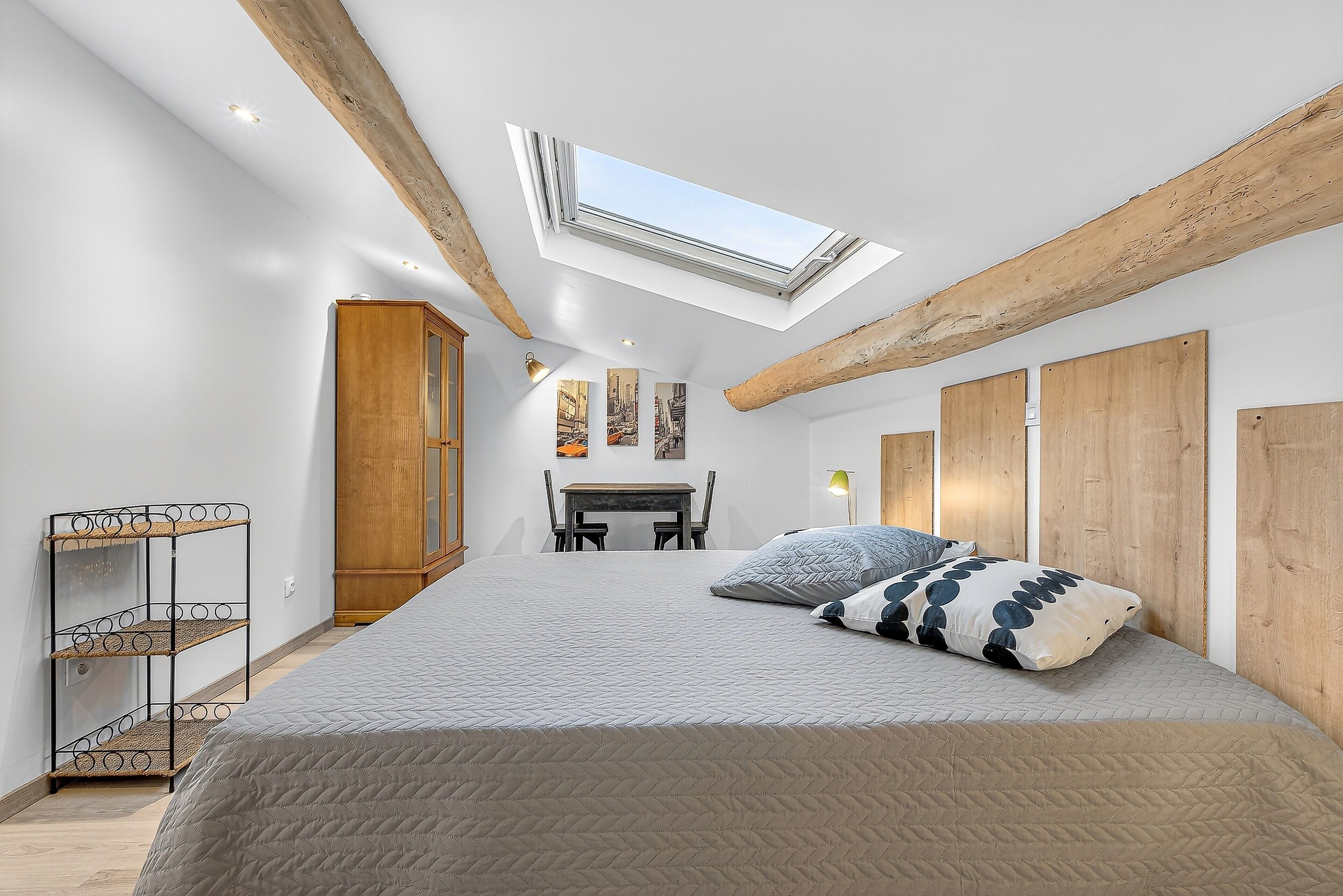 Un dormitorio con tragaluces, una cama con almohadones estampados, un armario de madera y una pequeña mesa con sillas.