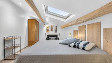 Un dormitorio con tragaluces, una cama con almohadones estampados, un armario de madera y una pequeña mesa con sillas.