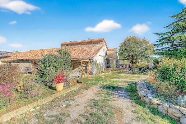 A rustic house with a tiled roof, surrounded by a garden with various plants and a stone wall.