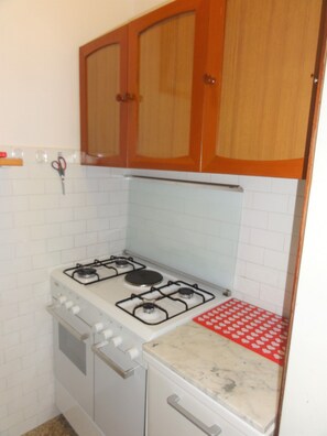A kitchen with white tiled walls, a stove with four burners, and wooden cabinets above. - Modern Coastal Apartment (San Michele al Tagliamento)