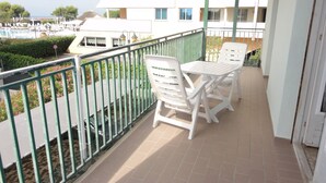A balcony with white chairs and a table, a green railing, and a view of a pool and buildings in the background.