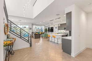Una cocina moderna con una isla central, un área de comedor con taburetes de bar, una escalera y una sala de estar con sofá y televisor.