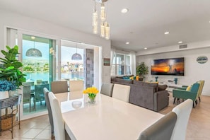 Una sala moderna con sofá, mesa de comedor y sillas, televisor montado en la pared y un amplio ventanal con vistas a edificios y una piscina.