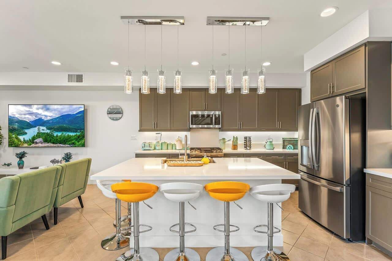 Une cuisine moderne dotée d’un îlot central, d’appareils électroménagers en acier inoxydable, de luminaires suspendus et d’un grand téléviseur à écran plat.