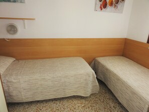 Una habitación pequeña con dos camas individuales, un cabecero de madera, un estante pequeño y una lámpara fijada a la pared.