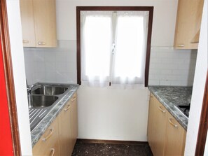 Una pequeña cocina con dos fregaderos, armarios de madera y una ventana con cortina blanca.
