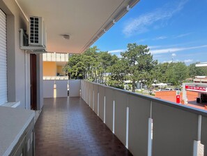 A covered balcony with a brick floor, white railings, and a view of a commercial area with a supermarket. - Apartment Near Shops and Beach (San Michele al Tagliamento)