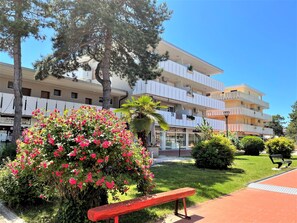 A multi-story hotel with balconies, a garden with pink flowers, and a red bench. - Apartment Near Shops and Beach (San Michele al Tagliamento)