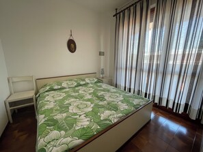 Un dormitorio con cama, una silla, una mesita de noche, un reloj de pared y una ventana con cortinas.