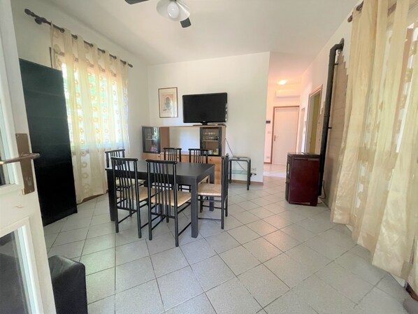 A dining area with a table and chairs, a television, and a kitchen area with cabinets and a refrigerator. - Bibione Pineda Villa With Private Garden (San Michele al Tagliamento)