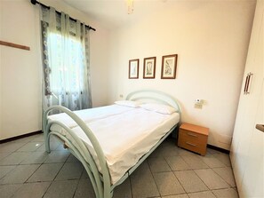 A bedroom with a bed, a nightstand, a window with curtains, and three framed pictures on the wall. - Bibione Pineda Villa With Private Garden (San Michele al Tagliamento)