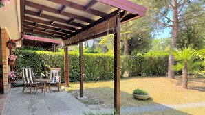 A covered patio area with a table and chairs, surrounded by greenery and a hedge. - Bibione Pineda Villa With Private Garden (San Michele al Tagliamento)