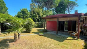 A small red building with a covered patio area, surrounded by a garden with various plants and trees. - Bibione Pineda Villa With Private Garden (San Michele al Tagliamento)