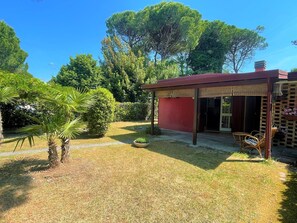 A small red building with a covered patio area, surrounded by a well-maintained garden with various plants and trees. - Bibione Pineda Villa With Private Garden (San Michele al Tagliamento)