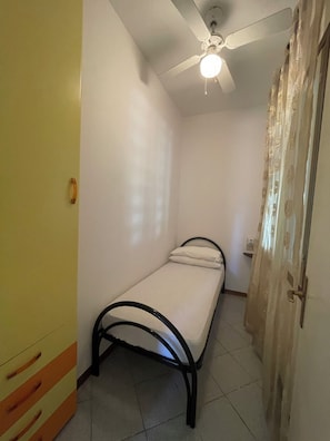 A small, single-bed room with a ceiling fan, a bedside table, and a curtain on the window. - Bibione Pineda Villa With Private Garden (San Michele al Tagliamento)