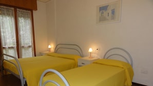 Una camera con due letti, ciascuno dotato di struttura in metallo e biancheria gialla. Sono presenti comodini con lampade e una finestra con tende in pizzo.