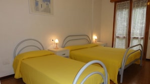 A room with two beds, each with a metal frame and yellow bedding. There are bedside tables with lamps, and a window with lace curtains.