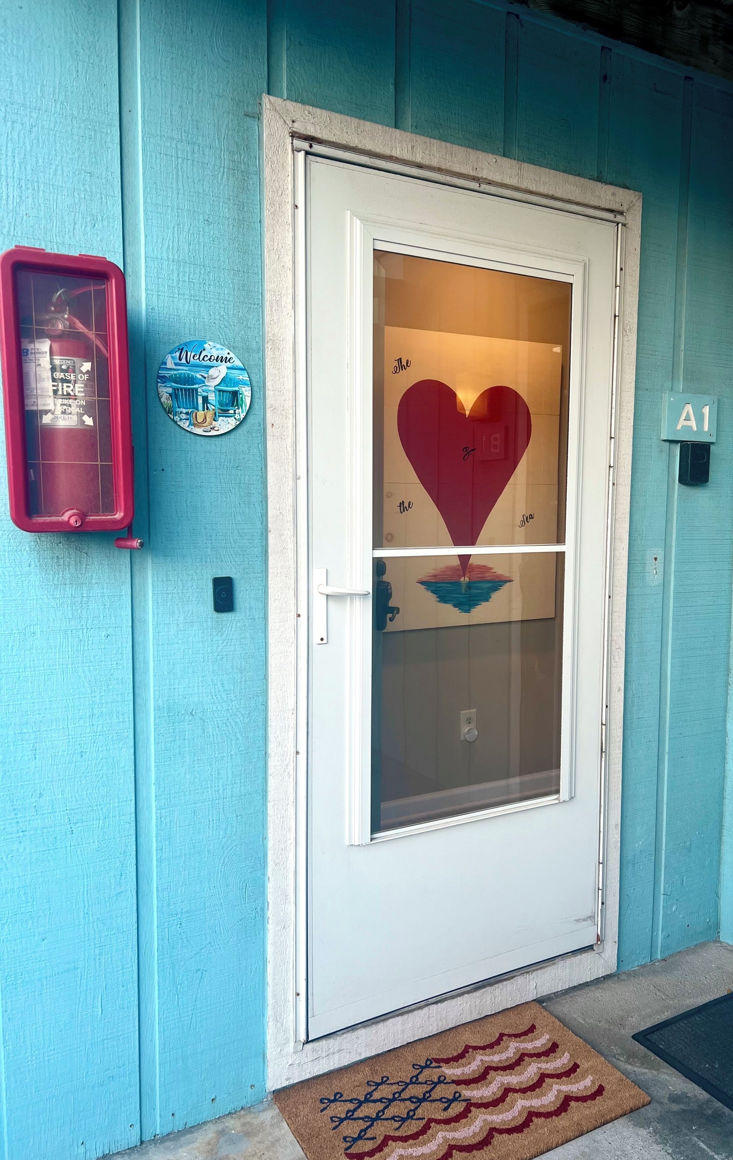 Una puerta con un ventanal en forma de corazón, un extintor, un letrero de bienvenida y un felpudo.