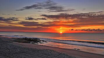 A beach at sunset with the sun near the horizon, calm sea, and a sandy shore.