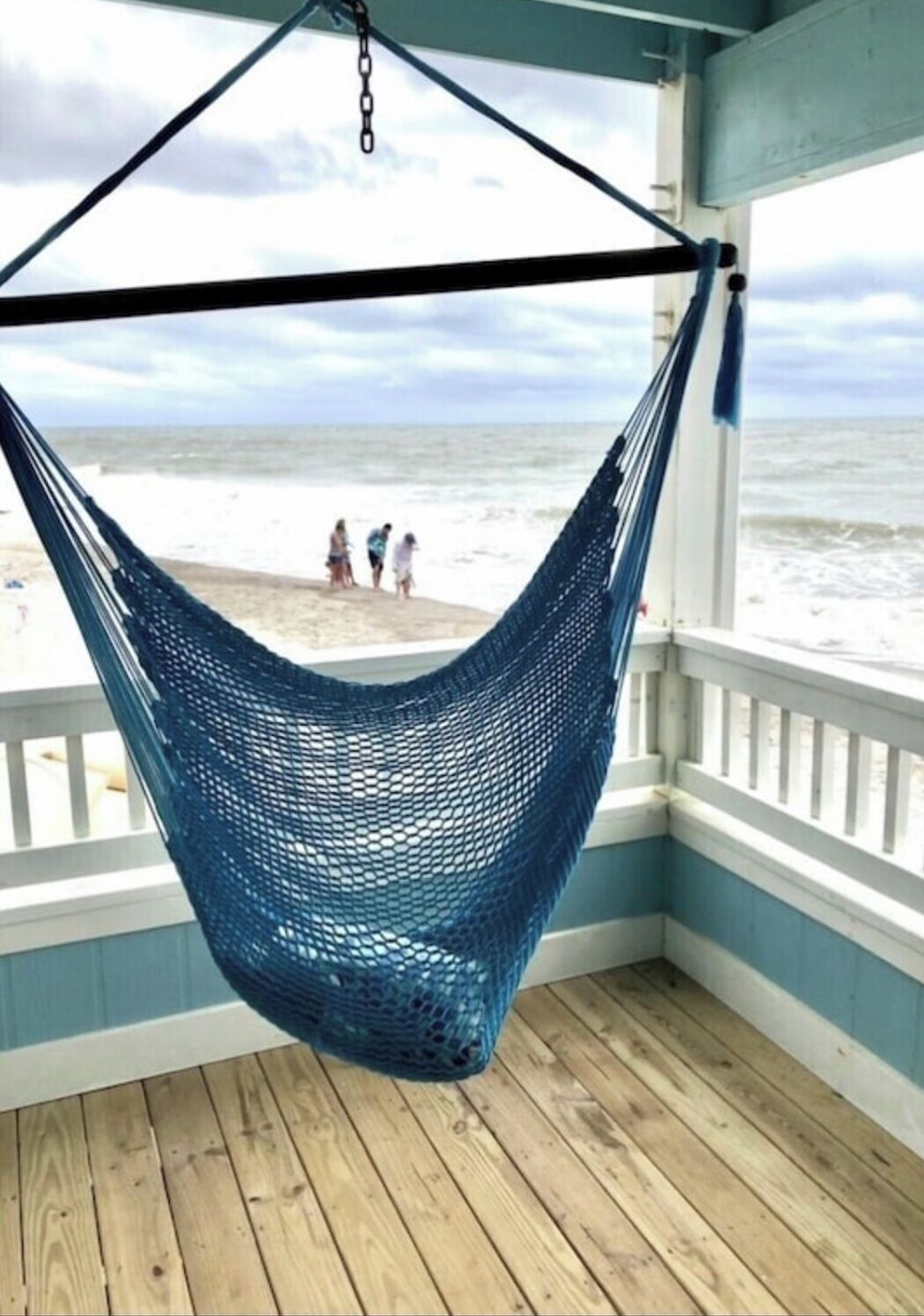 A blue hammock hanging from a ceiling in a room with a wooden floor and white railings, overlooking a beach with people in the distance.