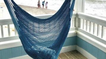 A blue hammock hanging from a ceiling in a room with a wooden floor and white railings, overlooking a beach with people in the distance.