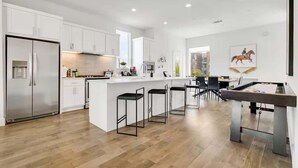 A modern kitchen with white cabinetry, a central island with bar stools, and a foosball table. - Designer Dallas Home, Rooftop, Kid Friendly Stay (Dallas)