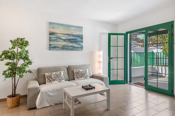 A living room with a sofa, a coffee table, a painting, a potted plant, and a sliding glass door leading to an outdoor area.