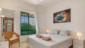 Un dormitorio con una cama grande, un ventanal verde, una silla de mimbre y un cuadro con una escena costera.