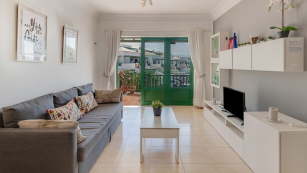 Un salón moderno con un sofá gris, una mesa de centro blanca, un televisor de pantalla plana y una puerta verde que da a un espacio exterior.